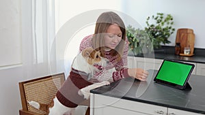 A dog in a brown sweater and a girl are sitting at a table with a tablet and candles. Jack Russell Terrier looks at