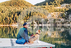 Dog owner and his friend beagle dog are sitting on the wooden pier on the mountain lake and enjoying the landscape during their