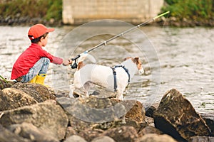 Dog watching his kid owner angling fish with fishing rod in river on summer day