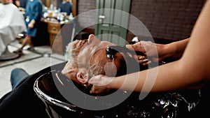 Before doing haircut. Handsome mature bearded man keeping eyes closed and relaxing while barber washing his hair in the