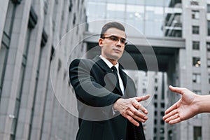 Doing handshake. Businessman in black suit and tie is outdoors in the city