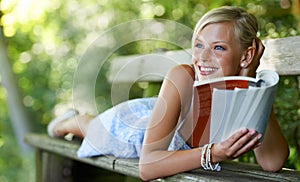 Doing some light reading. Pretty young woman lying on a bench and reading her book outdoors.