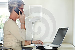 Doing some work from home. A senior woman talking on a cellphone while using her laptop at home.