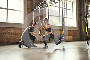 Doing squat exercise. Confident young personal trainer is showing slim athletic woman how to do squats with Trx fitness