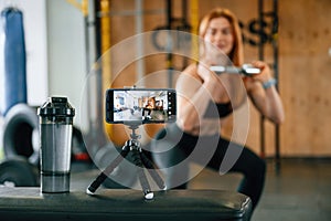 Doing squats, shooting video by smartphone. Beautiful strong woman is in the gym
