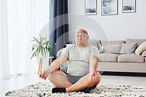 Doing yoga exercises. Funny overweight man in casual clothes is indoors at home