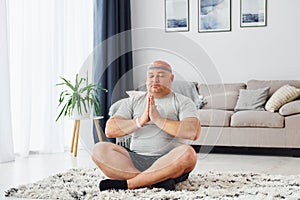 Doing yoga exercises. Funny overweight man in casual clothes is indoors at home