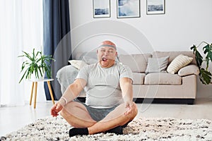 Doing yoga exercises. Funny overweight man in casual clothes is indoors at home