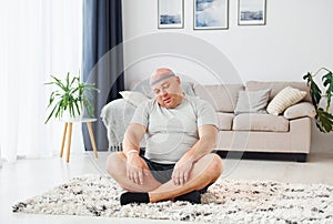 Doing yoga exercises. Funny overweight man in casual clothes is indoors at home