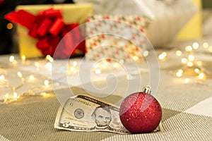 Dollar bill with red christmas decoration, gifts with lights and xmas tree on blurred background, festive greeting card