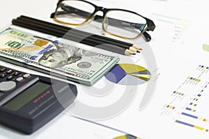 Dollar bills, calculator, pen, glasses, business charts are all on the table.