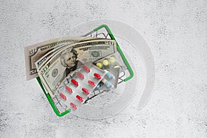 Dollars and pills in a mini grocery basket on a light background. Conceptual image of buying or selling drugs, healthcare, pharmac