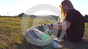 Dolly shot of beautiful girl sitting on green grass at field and stroking her siberian husky dog on sunset. Young woman
