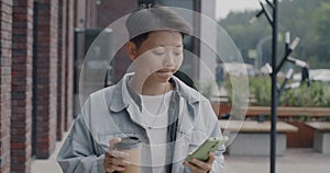 Dolly shot of joyful Asian man walking in city street using smartphone and drinking to go coffee