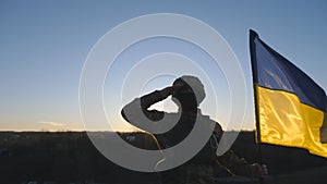 Dolly shot of male ukrainian army soldier with blue-yellow banner saluting against of sunset. Young military man in