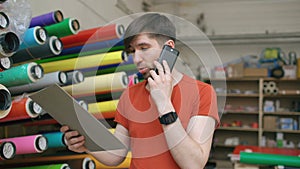 Dolly shot of smiling worker in warehouse talking smartphone and checking inventory. Man works in sales department of