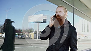 Dolly shot of Young bearded hipster man concentrated talking on phone on citystreet, conversation near office building