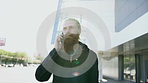 Dolly shot of young bearded hipster man concentrated talking on phone on citystreet, conversation near office building