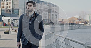 Dolly shot of young businessman walking along river embankment in city watching urban view