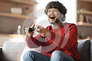 Domestic Fun. Cheerful Young Indian Guy Playing Video Games At Home