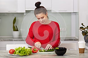 Domestic life and motherhood concept. Busy pregnant woman cuts vegetables for making salad, poses in modern apartment against