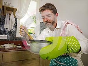 Domestic lifestyle portrait of happy and attractive  man in kitchen apron and glove holding cooking pot excited with delicious