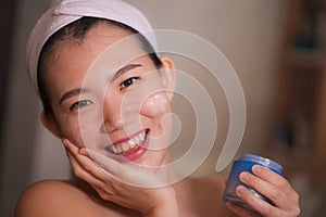 Domestic lifestyle portrait of young beautiful and happy Asian Chinese woman applying facial cosmetic cream smiling cheerful to
