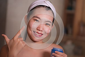 Domestic lifestyle portrait of young beautiful and happy Asian Korean woman applying facial cosmetic cream smiling cheerful to