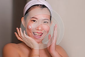 Domestic lifestyle portrait of young beautiful and happy Asian Korean woman applying facial cosmetic cream smiling cheerful to