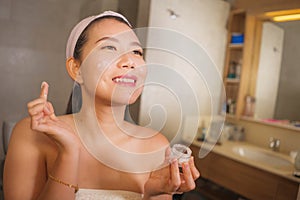 Domestic lifestyle portrait of young beautiful and happy Asian Korean woman applying facial cosmetic cream smiling playful to