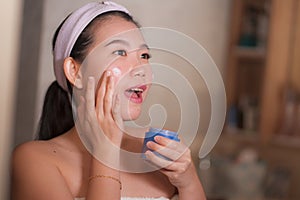 Domestic lifestyle portrait of young beautiful and happy Asian Korean woman applying facial cosmetic cream smiling playful to