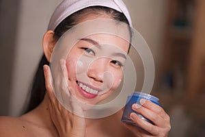 Domestic lifestyle portrait of young beautiful and happy Asian Korean woman applying facial cosmetic cream smiling playful to