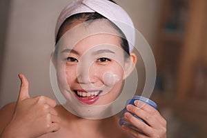 Domestic lifestyle portrait of young beautiful and happy Asian Korean woman applying facial cosmetic cream smiling playful to