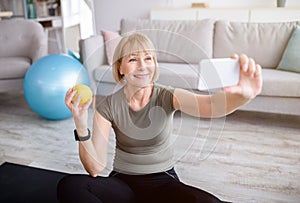 Domestic sports and healthy diet concept. Cheerful mature woman taking selfie with apple after home workout, indoors