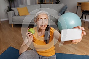 Domestic sports and healthy diet. Senior woman taking selfie with apple, resting after home workout on fitness mat