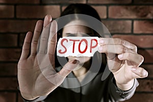 Domestic violence concept. Woman holding paper with word Stop near brick wall, focus on hands