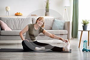 Domestic workout. Cheerful senior lady stretching her leg on sports mat in living room, empty space