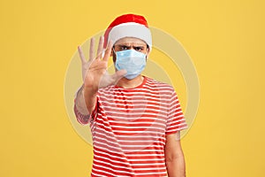 Don`t come to me and keep distance! Concerned man in medical face mask and santa claus hat holding hand showing stop gesture