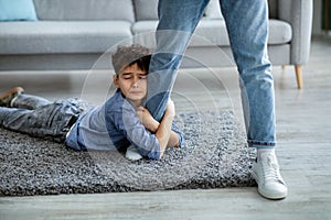 Don't go, dad. Upset boy embracing his father leg, begging daddy to stay with him, lying on the floor carpet