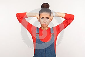 Don`t want to hear! Portrait of irritated girl with hair bun in denim overalls desperately closing her ears from loud noise