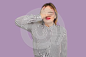 Don`t want to look at this! Portrait of shy confused woman with red lips in striped blouse covering eyes with hand