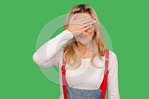 Don`t want to watch! Portrait of confused scared adult woman in stylish denim overalls covering eyes with hand