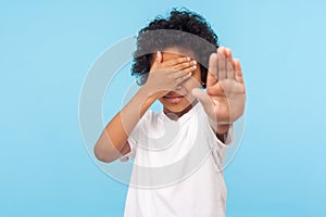 Don`t want to watch! Portrait of scared little boy with curls covering eyes with hand and showing stop gesture