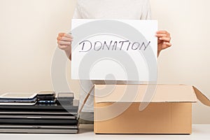 Donate technology to charity. Old used laptop computers, phone, tablet near cardboard box. Volunteer hands holding paper