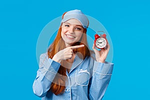 Dont forget wake up on time. Cheerful and cute relaxed feminine redhead woman in pyjama and sleep mask, holding red
