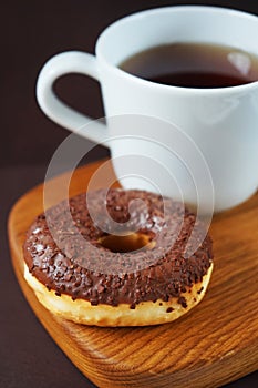 Donut with chocolate icing next to a white cup of tea on a wooden board