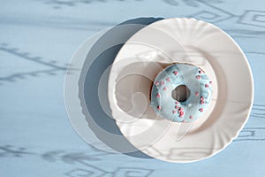 Donut covered with blue glaze and sprinkled with small pink hearts on a white plate on blue background under natural light