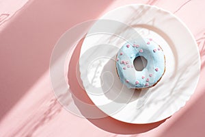 Donut covered with blue glaze and sprinkled with small pink hearts on a white plate on a pink background under natural light
