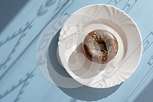 Donut covered with chocolate glaze and sprinkled with green tubes on white plate on blue background