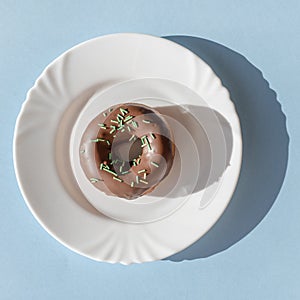 Donut covered with chocolate glaze and sprinkled with green tubes on white plate on blue background